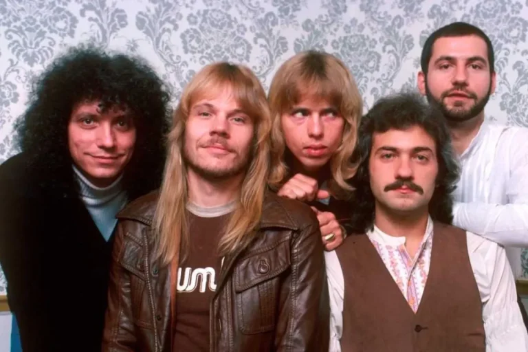 Styx band members posing together in a color studio portrait, wearing 1970s rock-era clothing with long hair, leather jackets, and patterned shirts against a decorative backdrop.