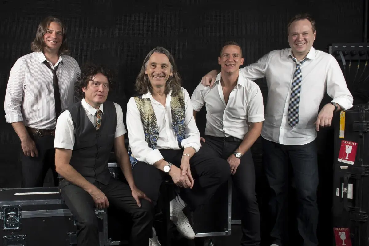 Modern lineup of Supertramp members smiling backstage with equipment cases.