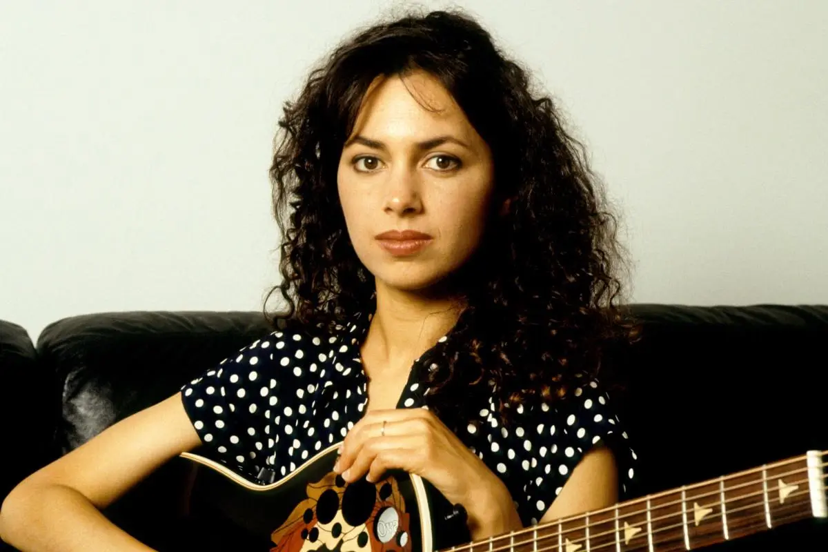 Portrait of Susanna Hoffs seated indoors, holding an acoustic guitar and looking directly at the camera with a calm expression.