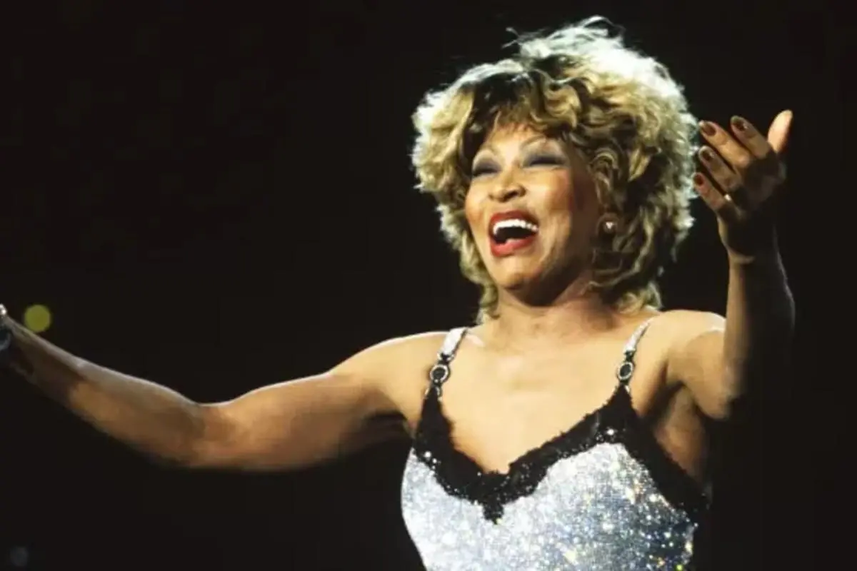 Tina Turner performs onstage with arms outstretched, smiling broadly under bright stage lights.