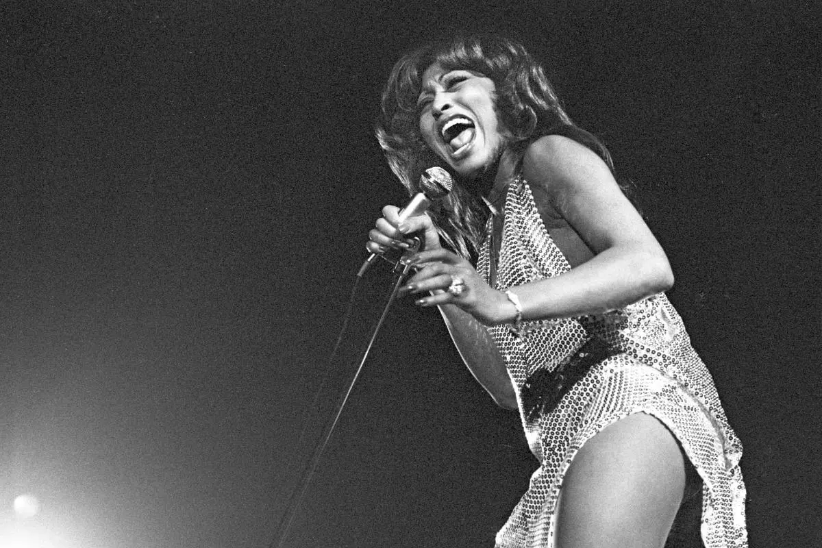 Tina Turner sings powerfully into a microphone, captured mid-note in a dynamic black-and-white performance shot.