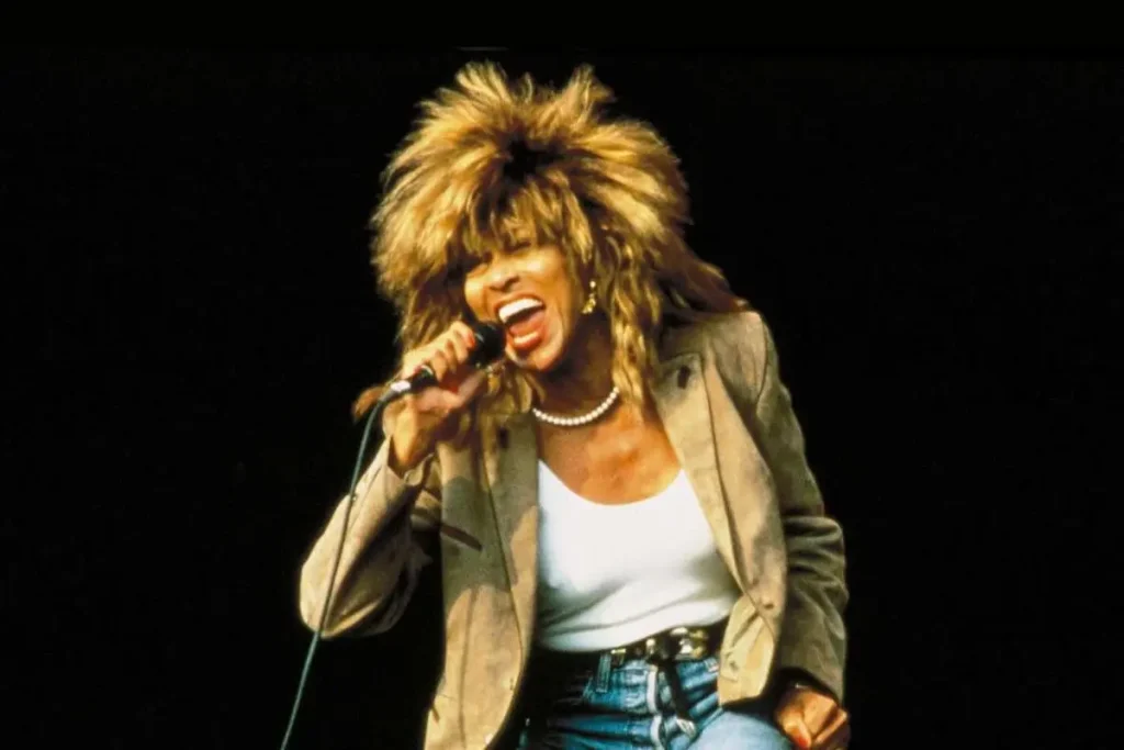 A high-energy stage performance shows Tina Turner leaning into the microphone, hair flying as she belts out a song.