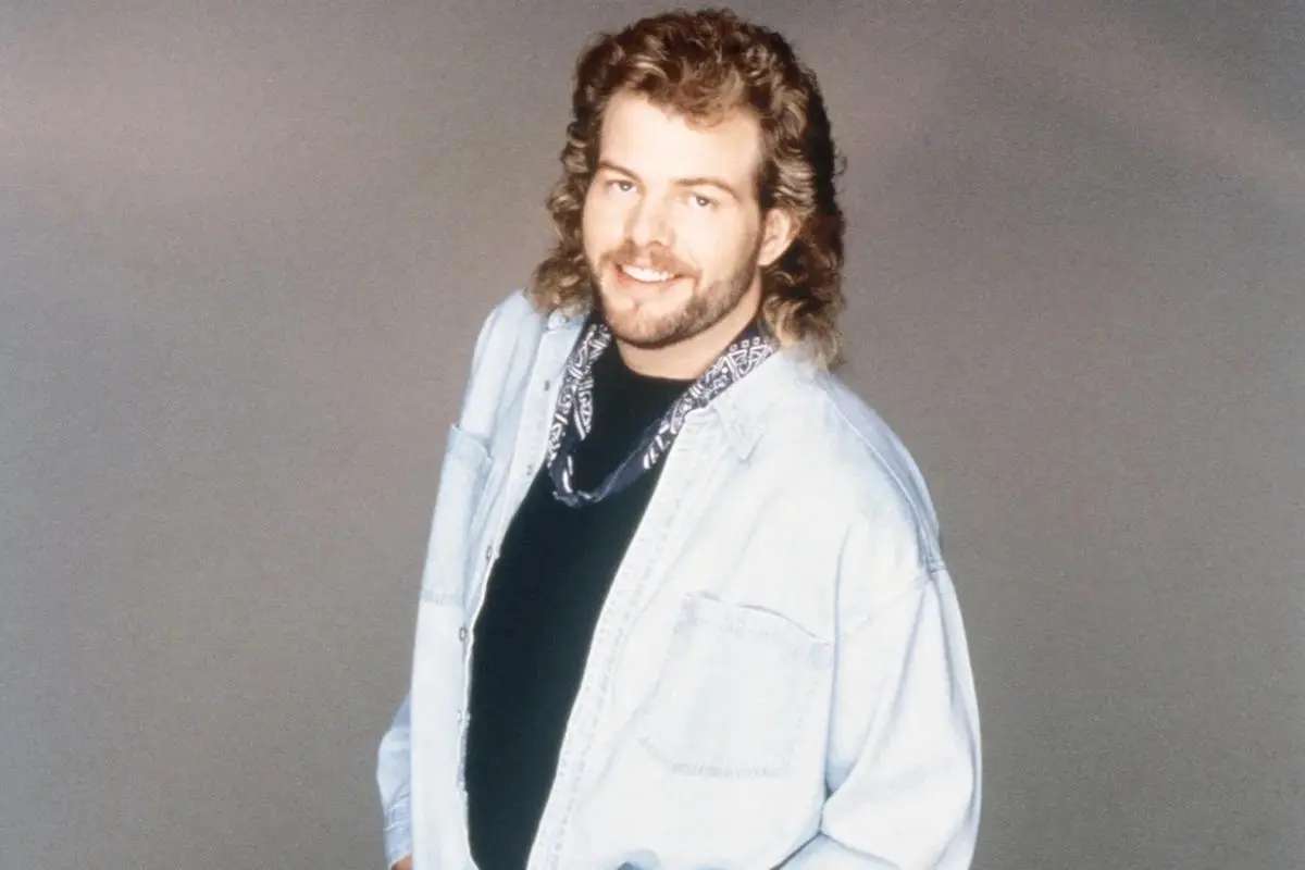 Toby Keith with a mullet hairstyle wears a light denim shirt and bandana against a plain background.