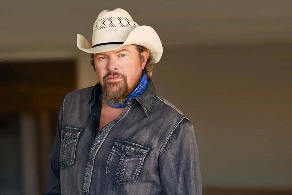 Toby Keith’s ‘Blame It on the Mistletoe’: Country Christmas for Grown-Ups Toby Keith in a white cowboy hat and denim shirt stands indoors, facing forward with a calm expression.