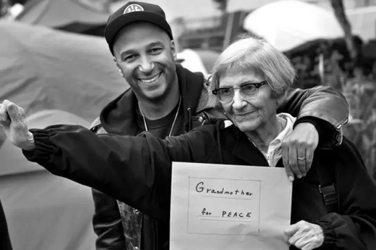 Maryn Morello holds a sign reading “Grandmother for Peace” while standing beside Tom Morello who has his arm around her during an outdoor gathering.