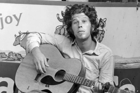 Tom Waits seated with an acoustic guitar, looking directly at the camera in a black-and-white portrait.