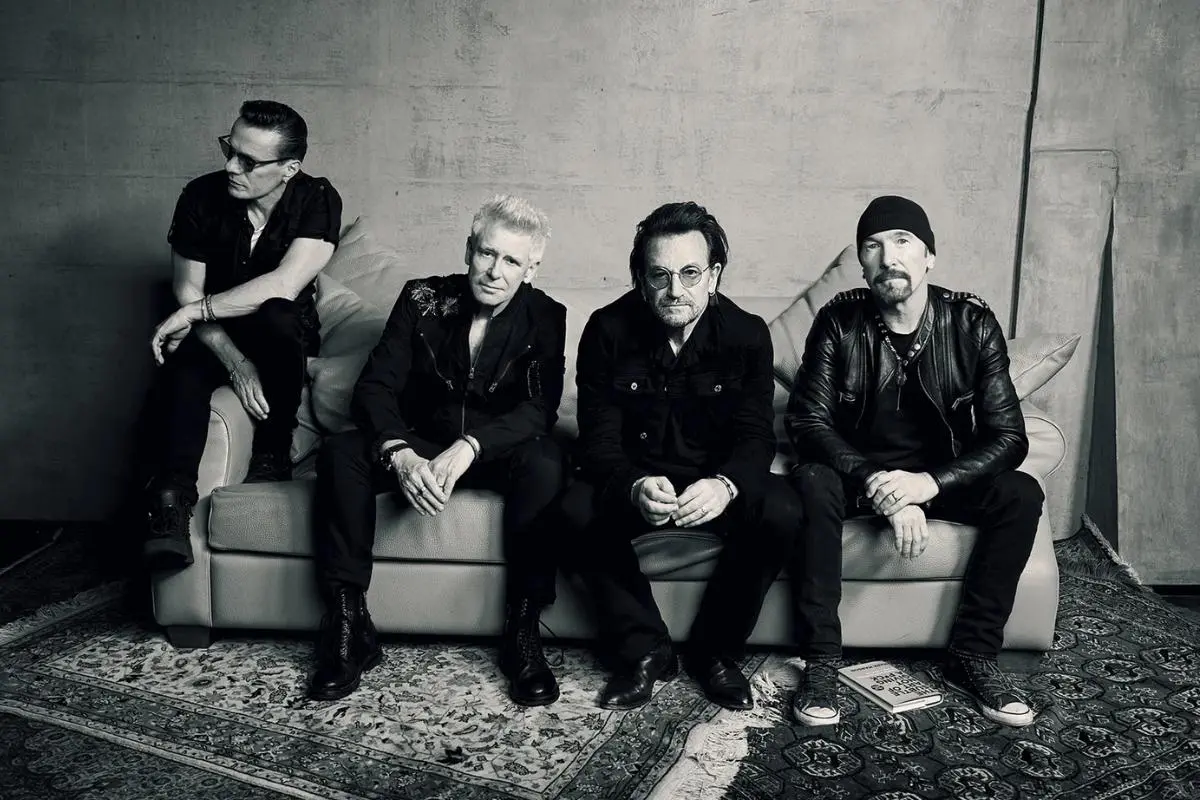 U2 seated on a couch, dressed in dark clothing, looking toward the camera.
