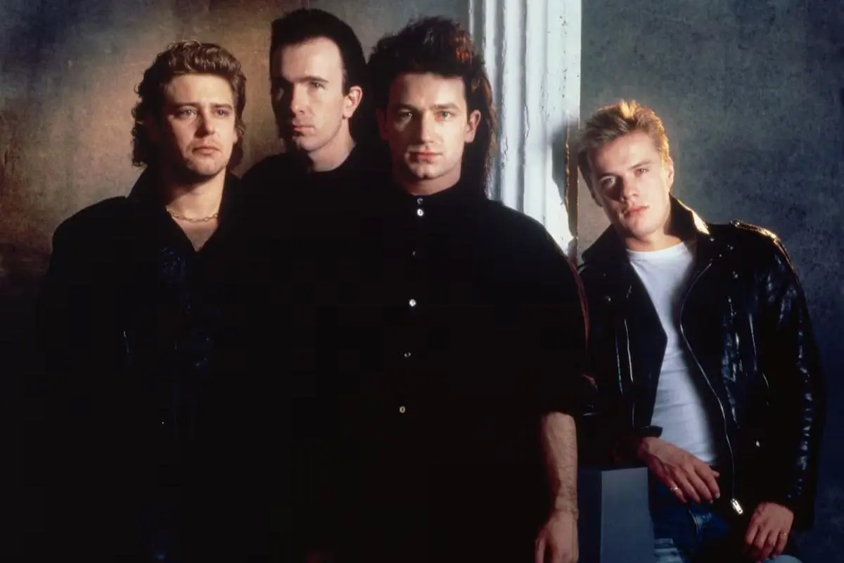 U2 band members standing close together indoors, wearing dark jackets and serious expressions.