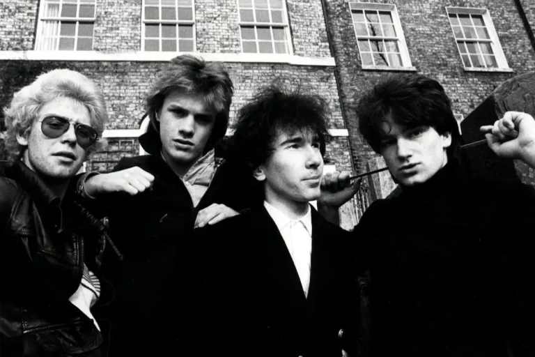 U2 band members outdoors, posing closely with intense expressions in front of brick buildings.