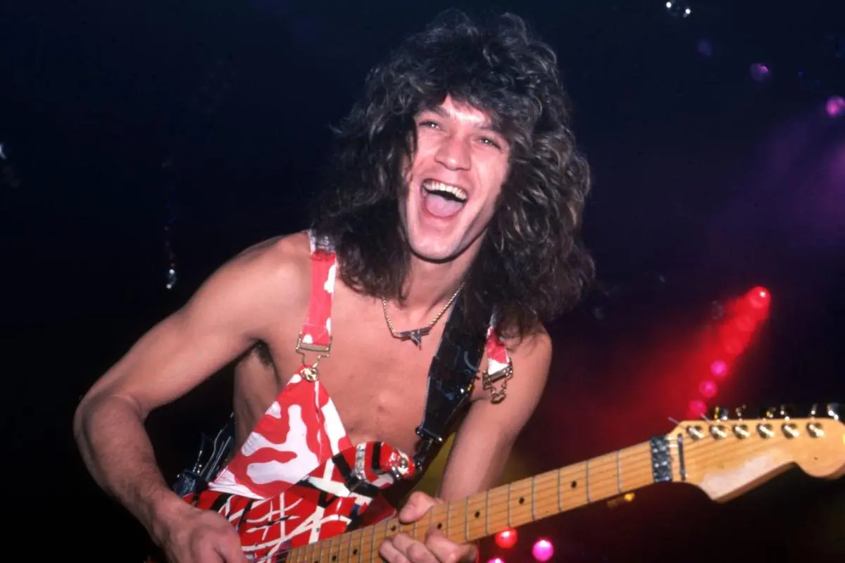 Van Halen smiles widely while playing a red-and-white striped electric guitar onstage.