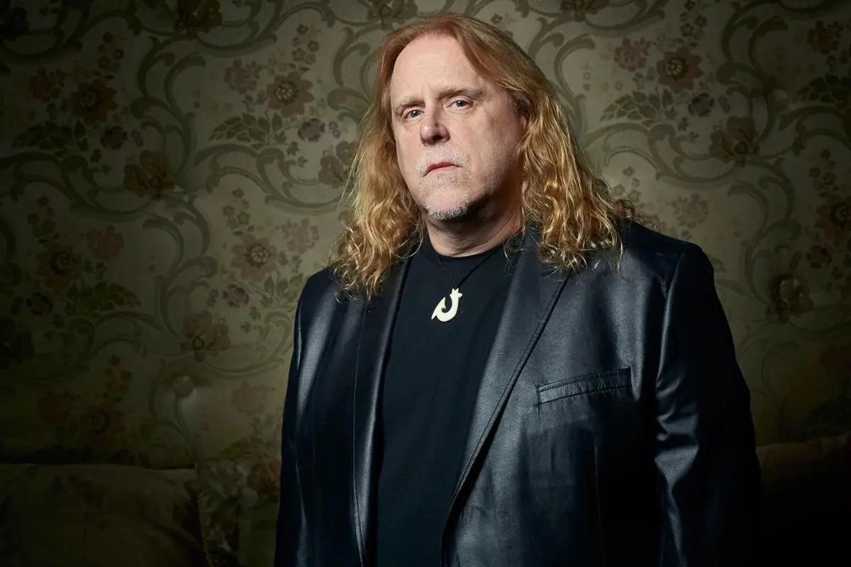 Warren Haynes in a studio portrait wearing a black jacket, standing against patterned wallpaper.