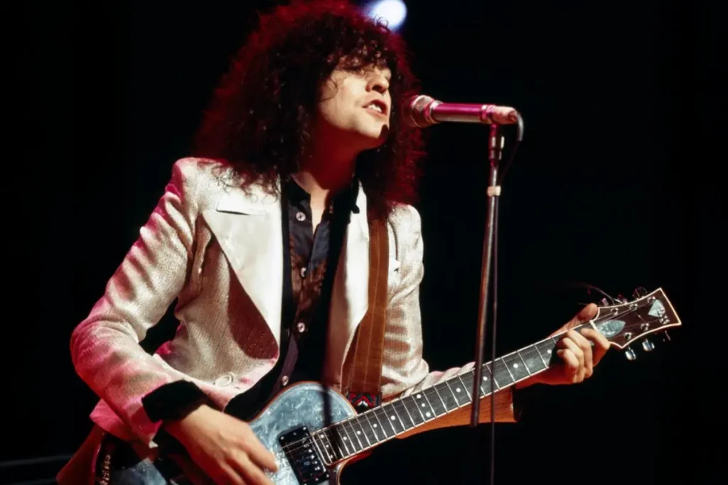 Marc Bolan sings into a microphone while playing an electric guitar and wearing a shiny silver jacket onstage.