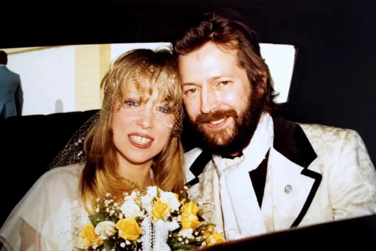 Pattie Boyd holding a bouquet of yellow flowers smiles beside Eric Clapton, both seated and dressed in formal wedding attire.