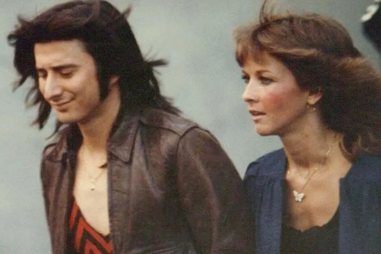 Steve Perry in a leather jacket walks beside Sherrie Swafford with windblown hair, both appearing deep in thought as they move forward together.
