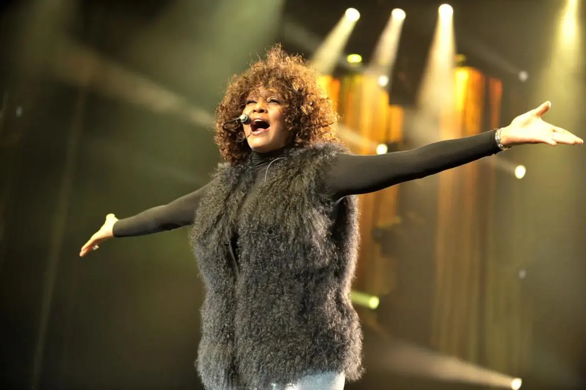 Whitney Houston performing live with arms outstretched, wearing a dark outfit under warm concert lighting.