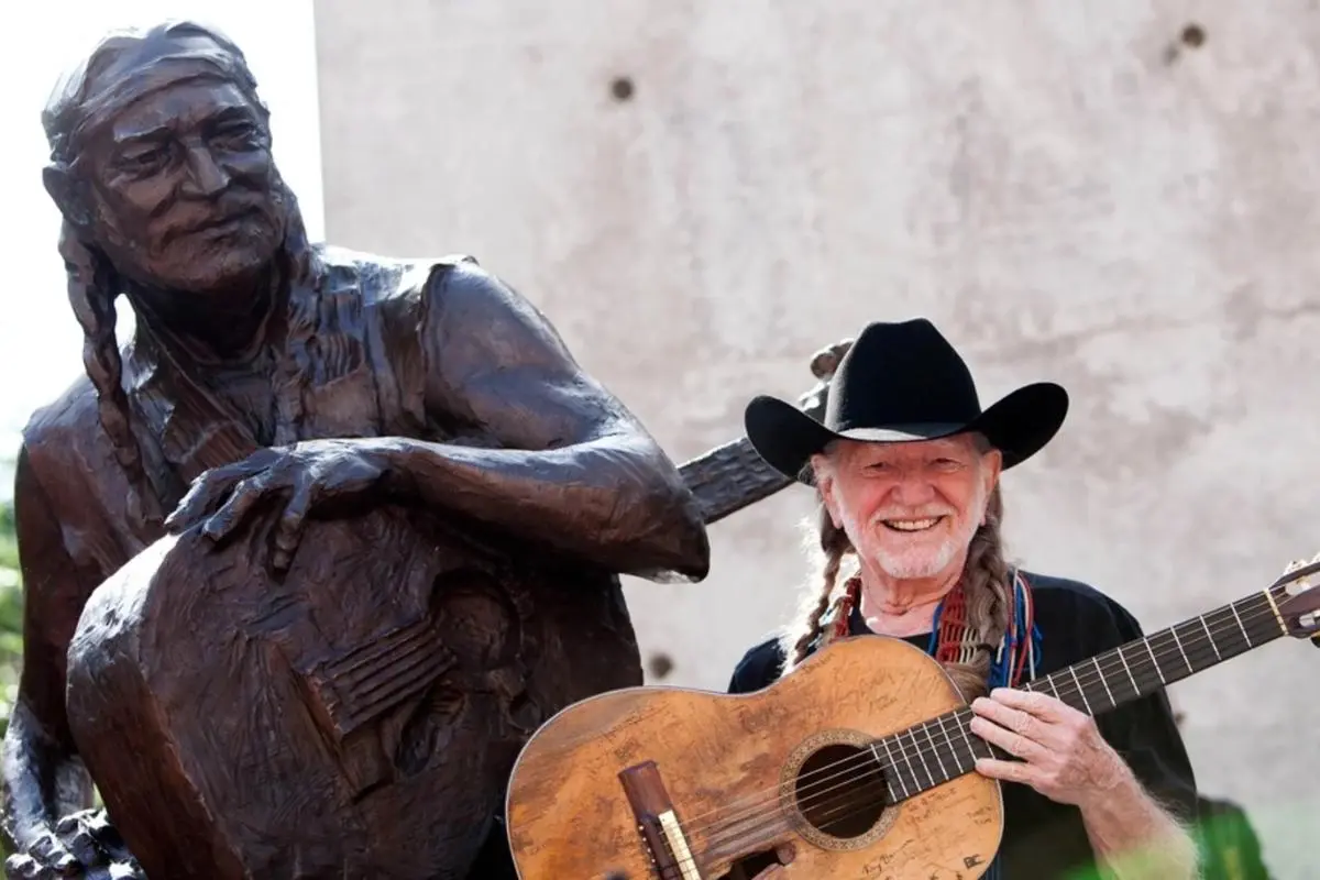 Willie Nelson smiling while holding an acoustic guitar beside a bronze statue, wearing a black cowboy hat.