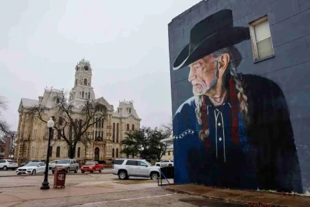 Large outdoor mural of Willie Nelson wearing a black cowboy hat and braids painted on the side of a building, with a historic courthouse and parked cars visible across the street.