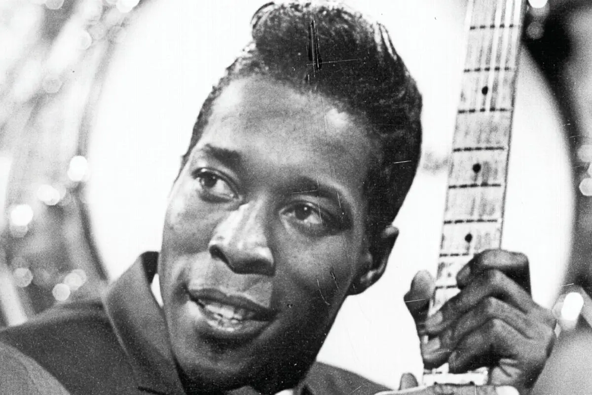 A young Buddy Guy in a suit holds the neck of his electric guitar close to his face, looking off to the side with a thoughtful expression.