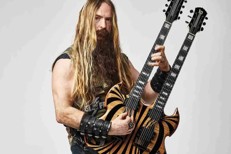 Zakk Wylde standing in a studio setting holding a double-neck electric guitar, wearing a sleeveless vest, leather wristbands, and long blond hair with a full beard.