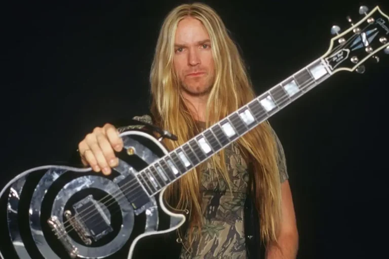 Zakk Wylde posing with a bullseye electric guitar against a dark background.
