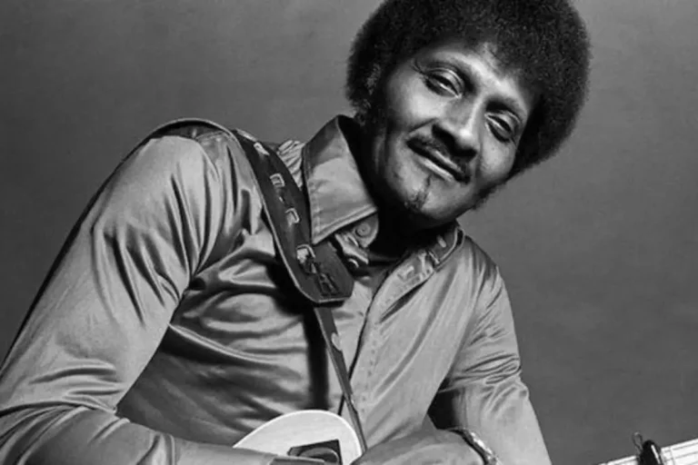 Albert Collins with an afro hairstyle poses with a guitar in a black-and-white studio portrait.