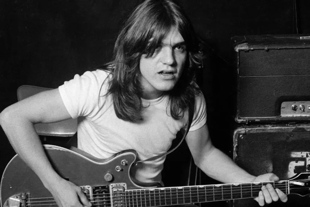 Angus Young Before AC/DC – Inside Kantuckee, the Lost 1972 Band Angus Young guitarist in a white T-shirt sits with an electric guitar, appearing mid-conversation or mid-song beside a stack of amplifiers.