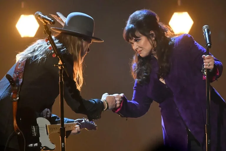 Ann Wilson and Jerry Cantrell onstage touch fists in a friendly gesture while standing at their microphones under warm lighting.