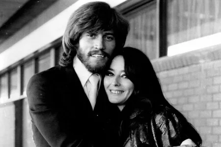 Young Barry Gibb with Linda Gray couple smiles while embracing closely, with the man’s arm wrapped around the woman as they stand outside a building.