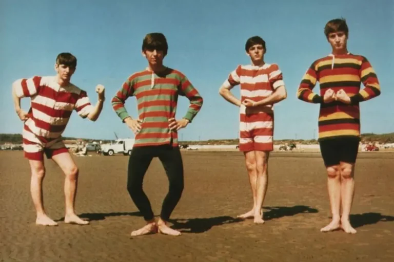The Beatles posing barefoot on a sandy beach in matching striped swimwear, standing in a line with playful, flexed poses under a clear blue sky.