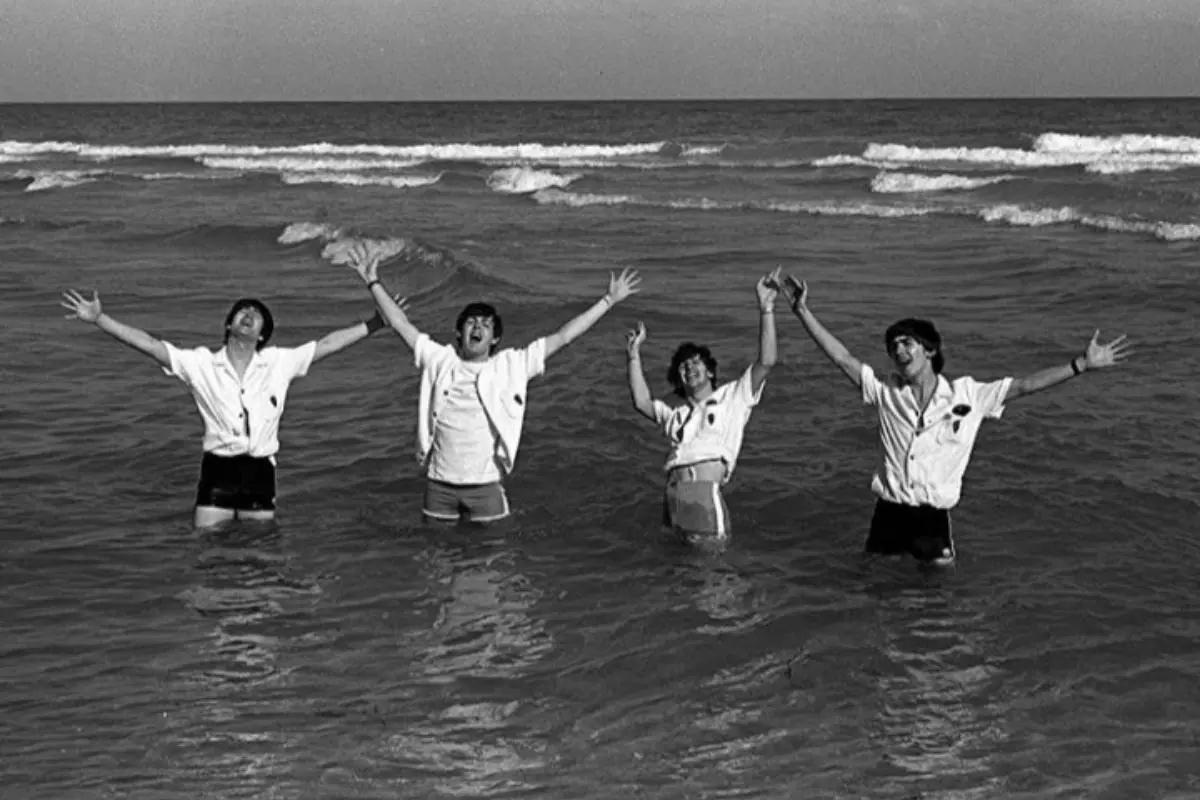 The Beatles standing waist-deep in the ocean in a black-and-white photo, raising their arms joyfully while wearing matching light-colored shirts during a seaside outing.