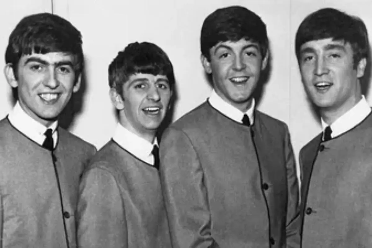 The Beatles posing together in a classic black-and-white portrait, wearing matching collarless suits and ties, smiling toward the camera in an early-career publicity photo.