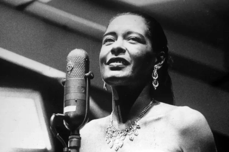 Billie Holiday singing into a vintage microphone while wearing elegant jewelry, black-and-white photo.