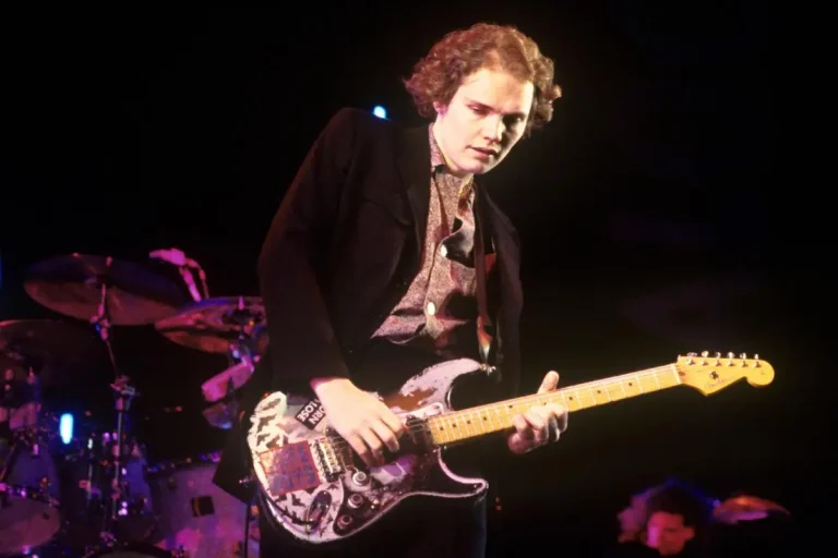 Billy Corgan playing electric guitar onstage under stage lights.