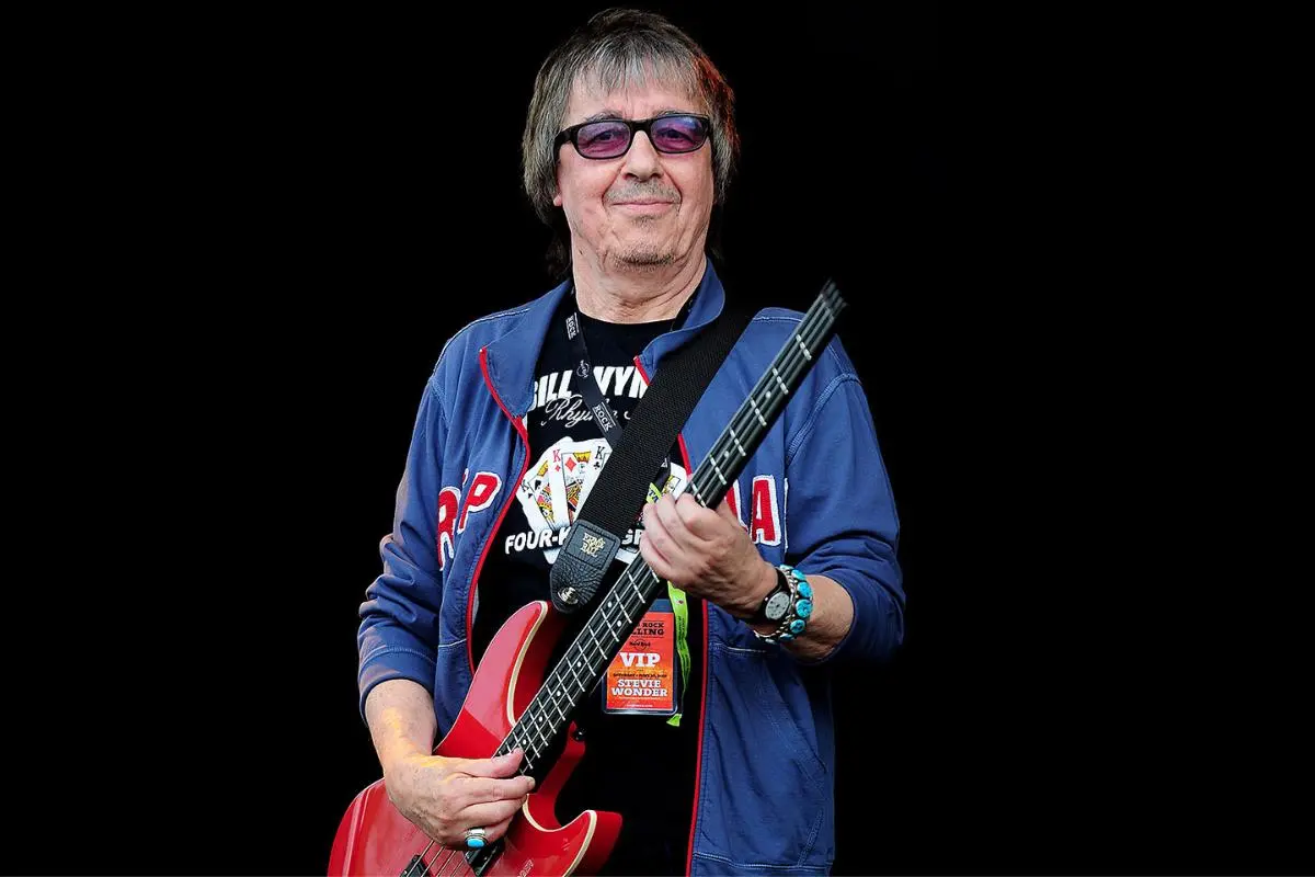 Bill Wyman stands against a black background holding a red electric bass.