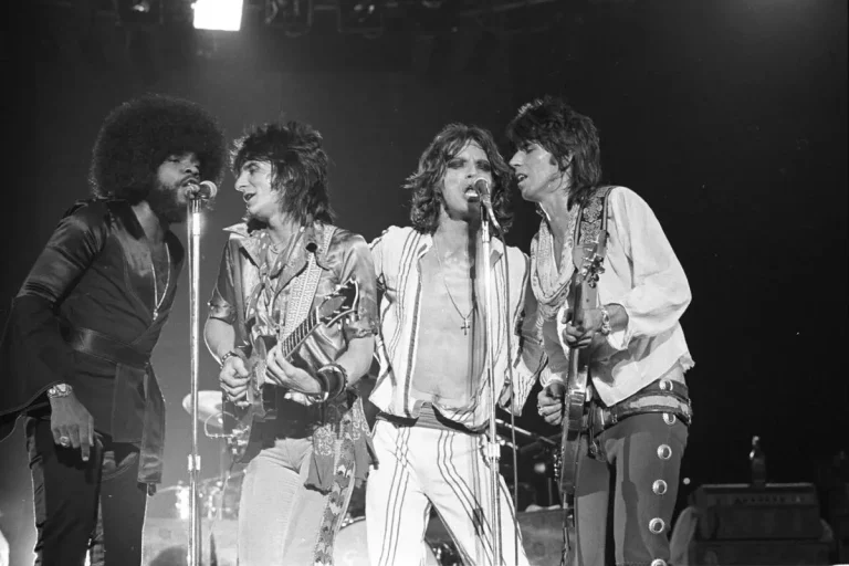 Billy Preston performing with the Rolling Stones singing together while two guitarists play beside them under bright concert lights.