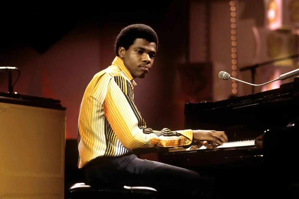 Billy Preston in a striped yellow shirt plays the piano onstage, glancing toward the camera as warm lighting glows behind him.