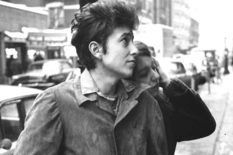 Bob Dylan looks upward while Suze Rotolo leans her head affectionately on his shoulder as they walk along a busy city street.