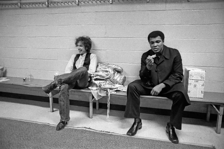 Bob Dylan and Muhammad Ali sitting on a bench in a sparse backstage room, one laughing with his legs crossed while the other looks down thoughtfully at something in his hand.