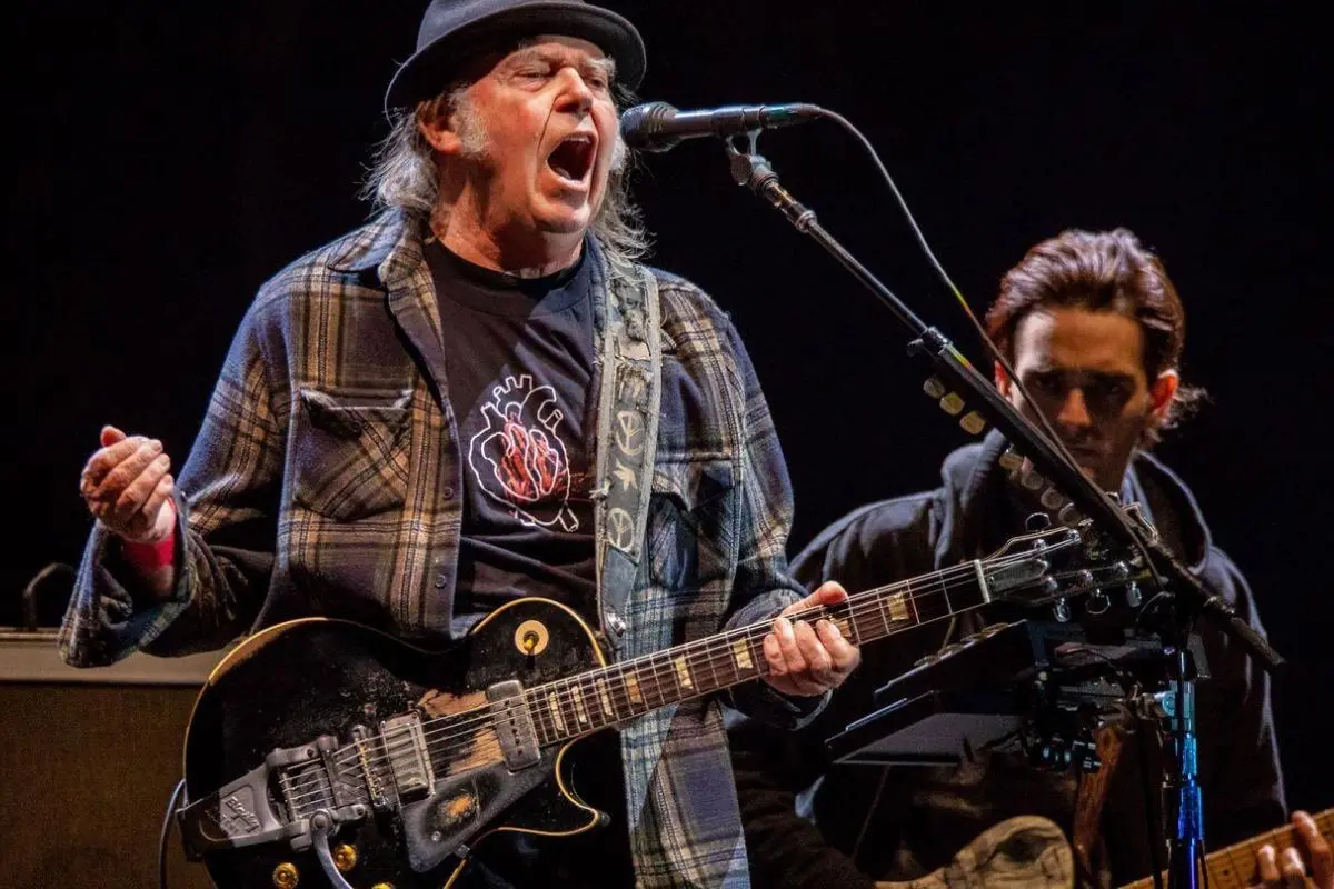 Neil Young performs onstage wearing a flannel shirt, black T-shirt, and a black hat.