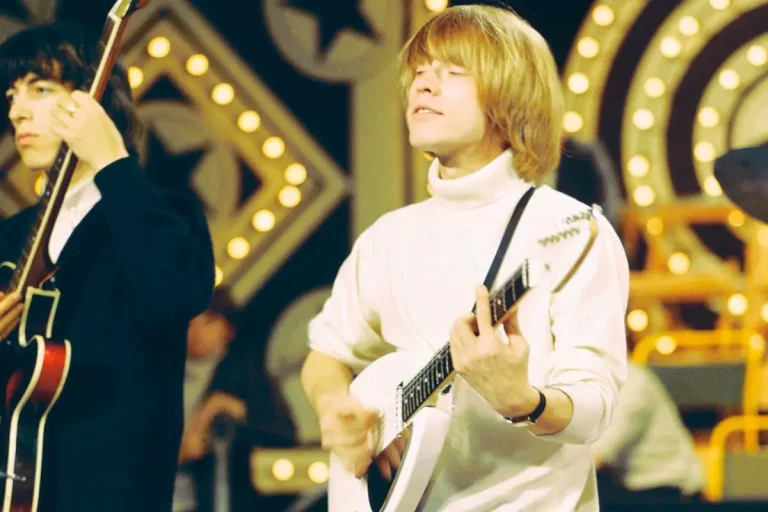 Brian Jones performing onstage, wearing a white turtleneck and playing a white electric guitar.