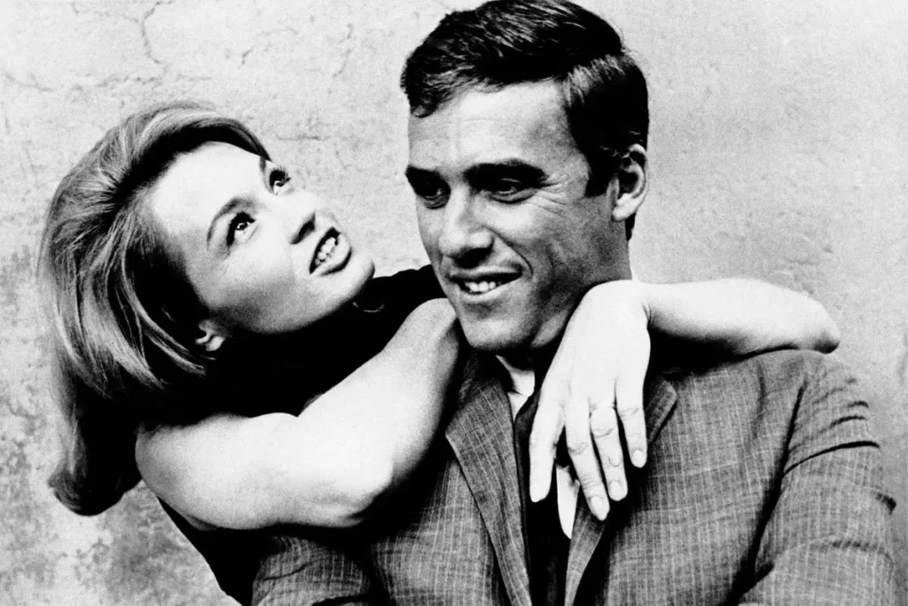 Burt Bacharach smiling as Angie Dickinson wraps her arms around his shoulders and looks up at him affectionately.