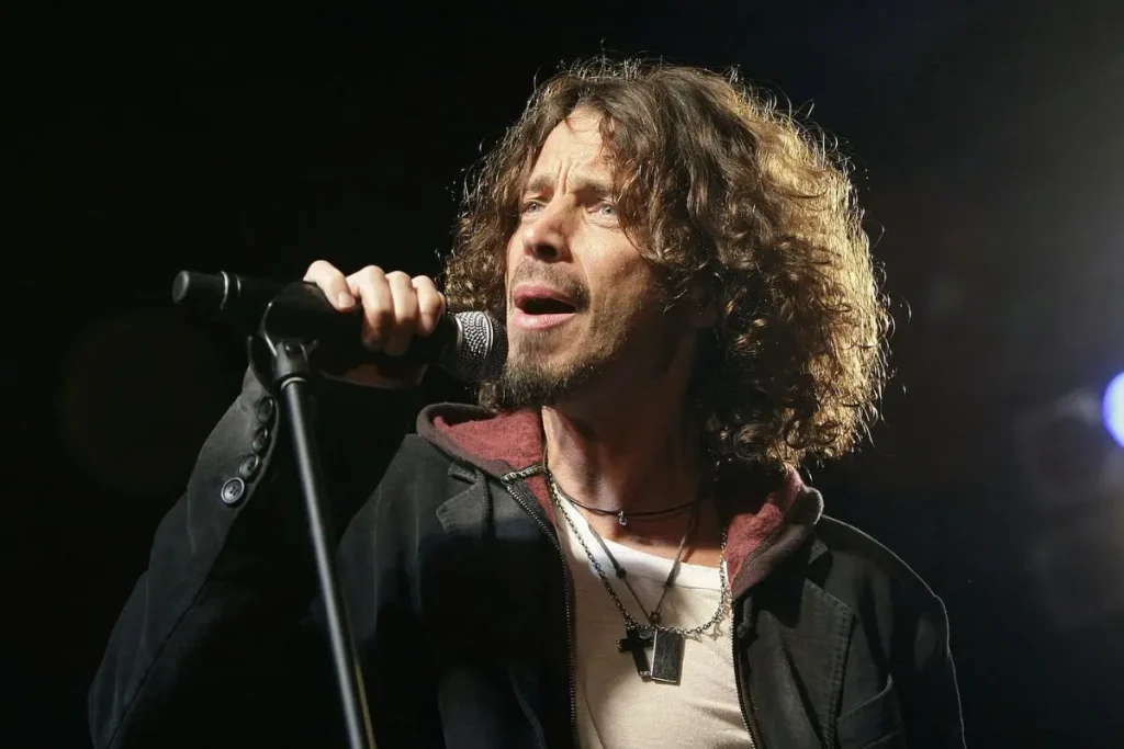 Chris Cornell holds a microphone and sings intensely onstage, wearing layered necklaces and a dark jacket.