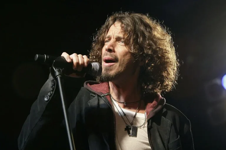 Chris Cornell holds a microphone and sings intensely onstage, wearing layered necklaces and a dark jacket.