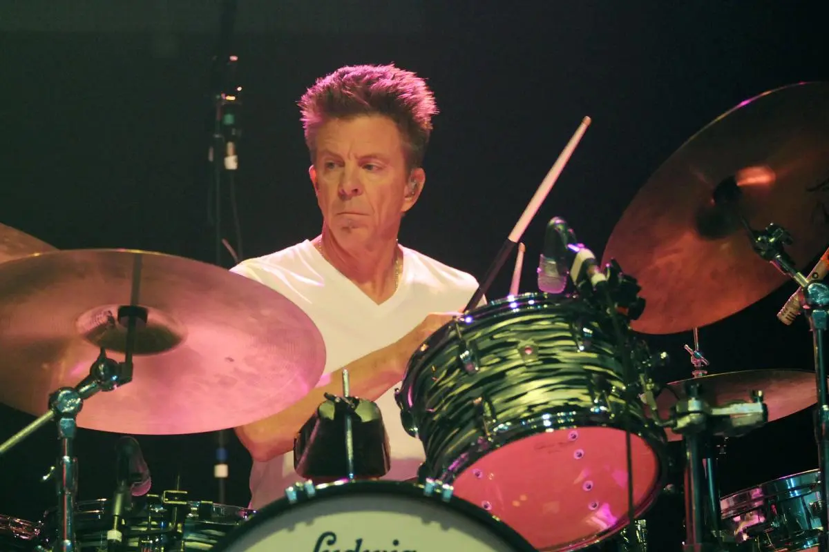 chris layton in a white T-shirt playing a drum kit onstage under warm lighting.
