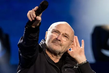 Phil Collins holding a microphone and cupping his ear during a live performance.