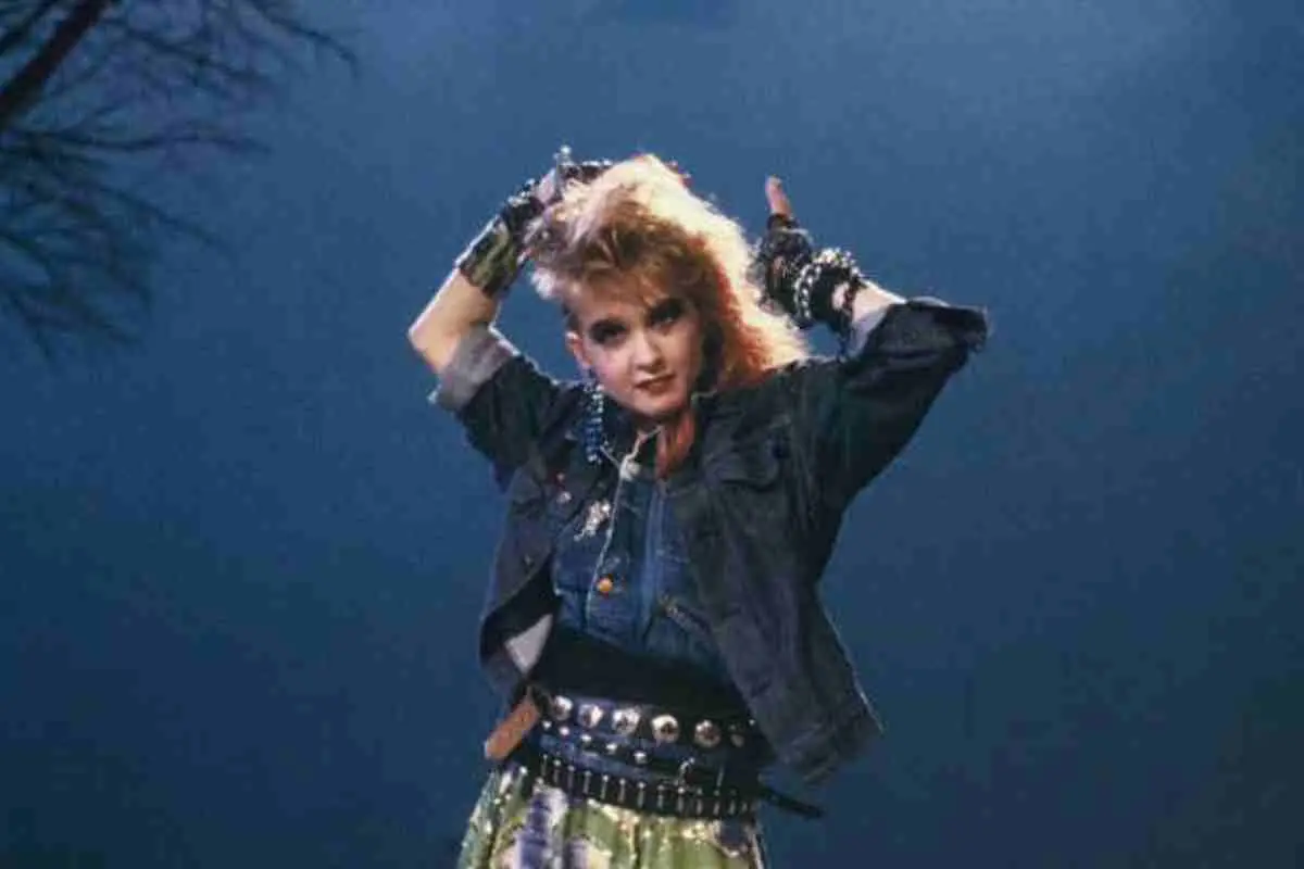 Cyndi Lauper in an early-career performance photo, standing on stage with arms raised, wearing layered 1980s punk-inspired fashion with denim and studded accessories.