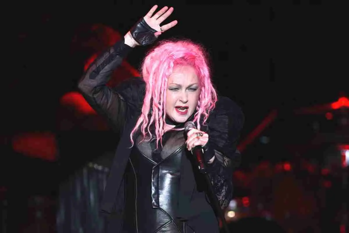 Cyndi Lauper performing on stage with bright pink hair, singing into a microphone with one hand raised, wearing a black outfit under dramatic concert lighting.