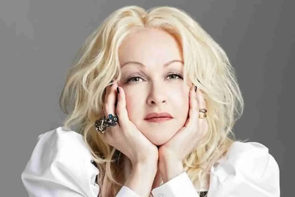 Cyndi Lauper posing in a studio portrait with light blond hair, resting her face in her hands, wearing a white top and statement rings against a neutral background.