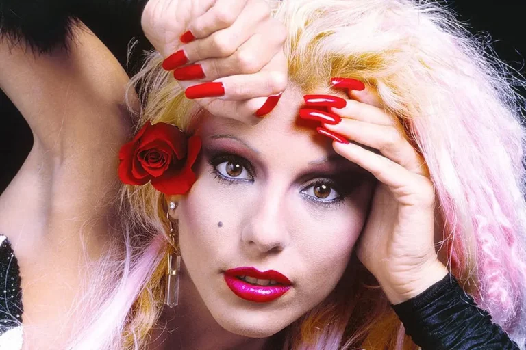 Dale Bozzio in a dramatic close-up, wearing bright red lipstick and long red nails, with a red rose tucked behind her ear.