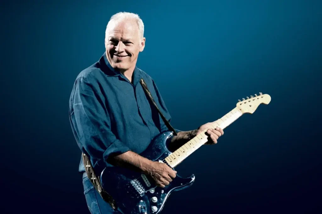 David Gilmour smiles while playing a dark blue Fender Stratocaster onstage.